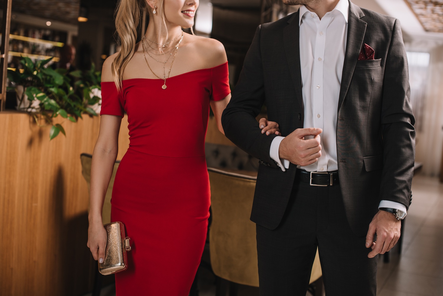 cropped view of boyfriend and girlfriend in red dress walking in restaurant and holding hands
